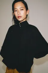 Camisa de algodón color negro, con cuello subido, manga larga terminada en puño ancho y bajo ajustable con cordones. Cuenta con cierre delantero con botones ocultos por solapa y detalles fruncidos.