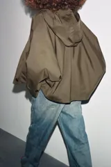 Trench corto color verde militar, con cuello solapa y capucha. Tiene manga larga con trabillas y botón en los puños, bolsillos delanteros y bajo asimétrico. Cierre frontal con botones. El tejido es repelente al agua.