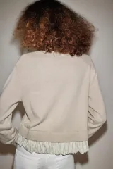 Chaqueta de punto color beige con cuello redondo, manga larga y cierre frontal con botones. Presenta un diseño combinado con volados de tela en el bajo.