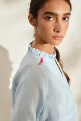Blusa de lino color marrón con escote en V, cuello con volados y mangas largas con puños fruncidos que presentan detalles en color crudo, al igual que en los hombros. Cuenta con cierre frontal de botones.