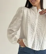 Blusa de brodery color blanco, con cuello redondo, mangas largas abullonadas con puños elásticos y cierre frontal con botones forrados. Presenta detalles de puntillas verticales en el frente.