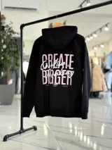 Canguro oversize de color beige claro con capucha, mangas largas y puños elastizados. Presenta un diseño gráfico en la espalda con el texto 'Creative Thinking' en tipografía cursiva negra sobre un fondo de letras grandes en tono rosado.
