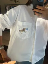 Camisa blanca de manga larga con cuello clásico y cierre frontal de botones. Presenta un bolsillo en el pecho con un bordado detallado de un caballo estilo carrusel, acompañado de estrellas y una estrella fugaz azul.