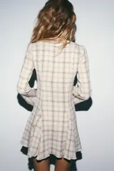 Vestido corto de algodón con estampado de cuadros en tonos beige y marrón. Presenta escote en pico, manga larga con corte evasé y bajo con volantes. Incluye cierre frontal con ganchos metálicos y cremallera lateral oculta.