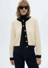 Chaqueta de punto de textura suave en color crema con detalles en contraste negro en cuello, puños, ruedo y tapeta de botones. Presenta un diseño recto, cuello redondo y cierre frontal con botones dorados.