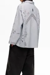 Camisa de manga larga color celeste, de corte holgado, con cuello clásico, cierre frontal de botones, bolsillo en el pecho y detalles de bordados florales y encaje en el ruedo y los puños.