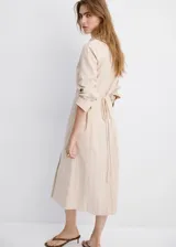 Vestido midi cruzado beige con sutiles rayas verticales oscuras, cuello camisero con solapa, mangas largas y cierre delantero cruzado.