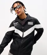 Campera deportiva de poliéster con diseño de bloques de color en negro y blanco. Presenta cuello alto, cierre frontal con cremallera y parches de la NASA y la bandera de Estados Unidos en el pecho.