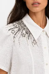 Vestido camisero midi de lino natural color blanco, con silueta fluida de línea A, cuello camisero, mangas cortas y un elaborado bordado de pedrería y cristales con diseño de estrellas en el hombro.