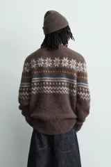 Jersey de punto con textura, corte relajado, cuello redondo y manga larga. Presenta un diseño jacquard geométrico con motivos de copos de nieve en tonos marrones, azules y blancos.