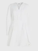 Vestido camisero corto color blanco, confeccionado en algodón orgánico. Presenta cuello en V, cierre frontal con botones, mangas largas abullonadas, cinturón de lazo ajustable en la cintura, bolsillos laterales y detalle de encaje calado en forma de escalera.