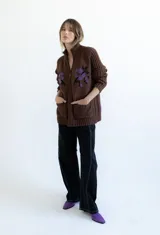 Cardigan de punto color marrón con cierre frontal, cuello alto y bolsillos aplicados. Presenta diseño tejido localizado con detalles en violeta y negro en el pecho.