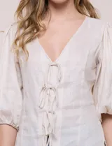 Blusa color beige confeccionada en lino, con escote en V pronunciado y cierre frontal de múltiples lazos para atar. Presenta mangas abullonadas tres cuartos con puños elásticos y corte relajado.
