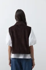Chaleco de pana color marrón oscuro, con cuello camisero, cierre frontal de botones metálicos dorados y dos bolsillos frontales con detalle de bordado geométrico.