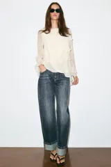 Blusa marrón de cuello redondo y manga larga, con volantes horizontales superpuestos y bajo asimétrico. Cuenta con detalle de abertura y botón en la espalda.