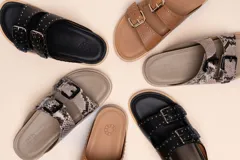 Sandalias de cuero color negro con dos tiras anchas sobre el empeine, cada una con hebillas metálicas ajustables y detalles de tachas plateadas. La suela es de cuero con un borde de color madera.