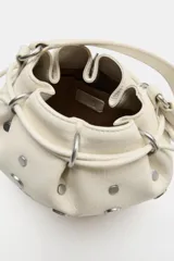 Bolso estilo saca de cuero color marfil, con diseño fruncido en la parte superior, tiras ajustables y tachas metálicas plateadas distribuidas por toda la superficie. Cuenta con una manija superior.
