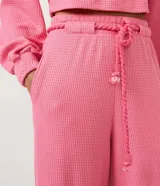 Pantalón tipo pantalona de tiro alto y corte amplio, confeccionado en tejido de ribana texturizada color rosado. Cuenta con cintura elástica y lazo ajustable con detalle de abalorios en los extremos.