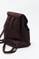 Mochila de símil gamuza color negro con textura tipo reptil. Presenta un diseño minimalista con solapa frontal, cierre de cordón ajustable y bolsillos laterales. Incluye un detalle metálico rectangular en la solapa.