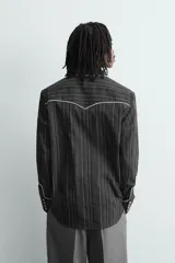 Camisa de corte relajado en color negro con rayas verticales finas. Presenta un diseño estilo western con vivos blancos en contraste en el canesú, cuello, tapeta frontal y puños. Cuenta con cuello solapa, cierre frontal de botones y mangas largas.
