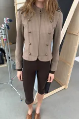 Blazer entallada color beige con rayas verticales finas, cuello redondo y manga larga. Presenta trabillas en los hombros y botones metálicos decorativos en el frente. Cierre frontal con cremallera y forro interior.