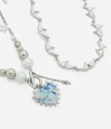 Collar doble de metal plateado compuesto por una cadena con eslabones ondulados y una segunda cadena con esferas metálicas, perlas, estrellas y un dije central en forma de corazón con acabado marmolado azul y blanco.
