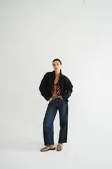 Campera de jean azul oscuro, estilo bomber, con cuello camisero y cierre frontal. Presenta un parche rectangular bordado en la espalda con patrón de rombos y texto.