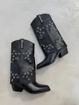 Botas texanas negras de caña alta, con diseño de pliegue superior y aplicación de múltiples ojalillos metálicos plateados en la caña y en la tira posterior. Poseen taco ancho y punta ligeramente cuadrada.