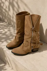 Botas texanas de gamuza color marrón, con caña alta, cierre frontal decorativo con tiras para anudar y hebilla lateral en el tobillo.