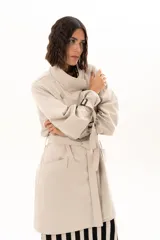 Trench corto color beige con cuello alto, cierre frontal de botones y cinturón ajustable en la cintura. Presenta mangas largas con detalles de tiras ajustables en los puños y bolsillos laterales.