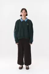 Buzo estilo rugby de corte oversized, color verde oscuro, con cuello tipo camisa en denim azul claro y cierre frontal parcial. Presenta un recorte horizontal en el pecho, logo bordado y tiras laterales en las mangas en contraste.