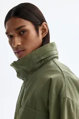 Campera estilo cargo de tejido ripstop, color verde militar, con cuello elevado que incluye capucha oculta. Presenta cierre frontal con solapa rompevientos, cuatro bolsillos delanteros con solapa y botón a presión, puños ajustables y cordón interno en la cintura.
