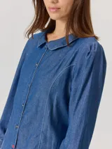 Camisa de denim con cuello bebé, cierre frontal con botones y costuras decorativas en contraste. Presenta mangas largas con sutil frunce en los hombros y un corte entallado.