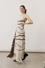 Vestido largo de punto sin mangas, con cuello redondo y ribete celeste. Presenta un diseño de líneas onduladas marrones sobre base cruda y un panel lateral con patrón geométrico y flecos en el bajo.