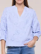 Blusa celeste a rayas con aplicaciones florales en relieve, escote en V y mangas 3/4 abullonadas con puños elásticos.