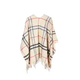 Poncho de tejido de punto con diseño de cuadros escoceses en tonos beige, negro y rojo. Presenta un corte amplio, cuello abierto y terminaciones con flecos en el ruedo.