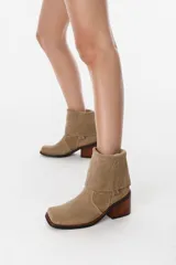 Bota de caña media confeccionada en cuero gamuzado color chocolate. Presenta un diseño de estilo western con taco cuadrado de 7 cm y una correa decorativa en el tobillo con herrajes metálicos plateados.