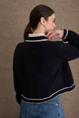 Cardigan de lana color marrón con detalles de bordes en color rojo. Presenta cierre frontal con botones, mangas largas con puños ajustados y un pequeño bordado decorativo en el pecho.