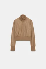 Chaqueta bomber de corte cropped confeccionada en material efecto piel color beige, con cuello subido, puños y cintura en rib elástico a tono, cierre frontal con cremallera y bolsillos laterales de vivo.