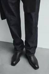 Botín de cuero negro con punta cuadrada, caña corta y cierre lateral con cremallera. Presenta una correa con hebilla metálica alrededor del tobillo.