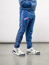 Pantalón deportivo de piqué color azul marino, con franjas laterales en contraste rojo y blanco. Presenta el escudo del Club Nacional de Football bordado en la pierna derecha y una etiqueta conmemorativa en la izquierda.