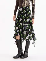 Pollera asimétrica de seda con estampado floral de rosas blancas sobre fondo negro. Presenta volados irregulares en forma de caracol que recorren el frente y la espalda, aportando volumen y movimiento.