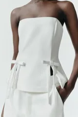Top strapless de satén color blanco, con diseño entallado y detalle de dos lazos decorativos en el frente. Presenta aberturas en el bajo y cierre posterior con cremallera.