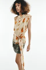 Vestido corto sin mangas de cuello redondo, con estampado tie-dye en tonos beige, naranja y verde oscuro. Presenta un lazo para atar en la cintura y cierre trasero con cremallera oculta.