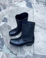 Botas de cuero vacuno color negro, con diseño versátil que incluye una polaina desmontable y un estribo decorativo con tachas metálicas. Presentan un taco bajo y punta redondeada.