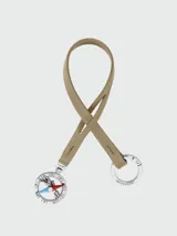 Llavero Longchamp modelo Le Pliage Baroudeur Compass, elaborado en cuero color tostado con una tira de diseño colgante. Cuenta con un aro metálico plateado y un dije colgante en forma de brújula con detalles esmaltados en rojo y azul y el logotipo de la marca.
