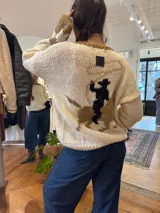 Buzo de tejido de punto artesanal en lana merino color crema, con diseño de estilo cowboy en la espalda que muestra una figura a caballo. Presenta detalles en color rojo en el cuello y en los puños de las mangas.
