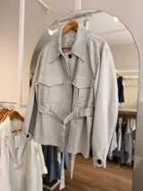 Campera blanca de algodón con cuello amplio, bolsillos delanteros con solapa, mangas largas y cinturón ajustable con hebilla redonda oscura. Cuenta con botones negros a contratono.