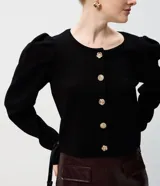 Cardigan de punto negro con cuello redondo, mangas largas abullonadas y cierre frontal con botones decorativos en forma de flor dorada. Presenta un corte corto y ajustado.
