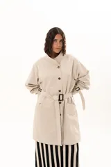 Trench corto color beige con cuello alto, cierre frontal de botones y cinturón ajustable en la cintura. Presenta mangas largas con detalles de tiras ajustables en los puños y bolsillos laterales.